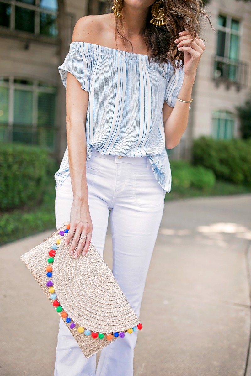 Amanda Miller wearing a white and blue off the shoulder top and white flare jeans and holding a straw pom clutch